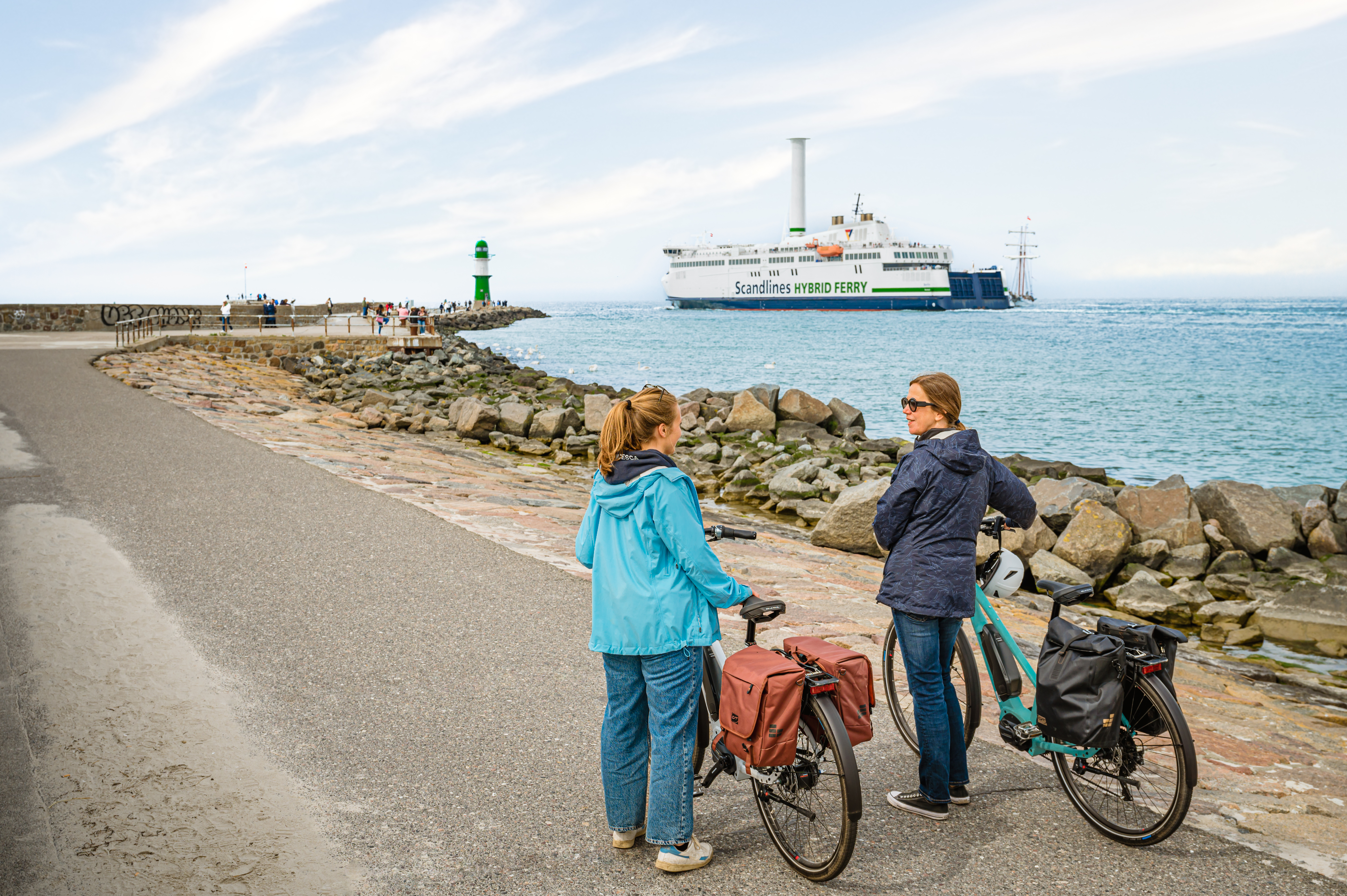 Zwei Frauen stehen mit dem Rücken zur Kamera und ihren Fahrrädern auf der Mole in Warnemünde. Auf der Ostsee fährt eine Fähre und im Hintergrund sieht man den grünen Leuchtturm